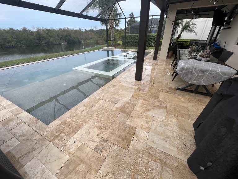Cleaned travertine pool deck surrounding a screened infinity pool with lakefront view in Florida